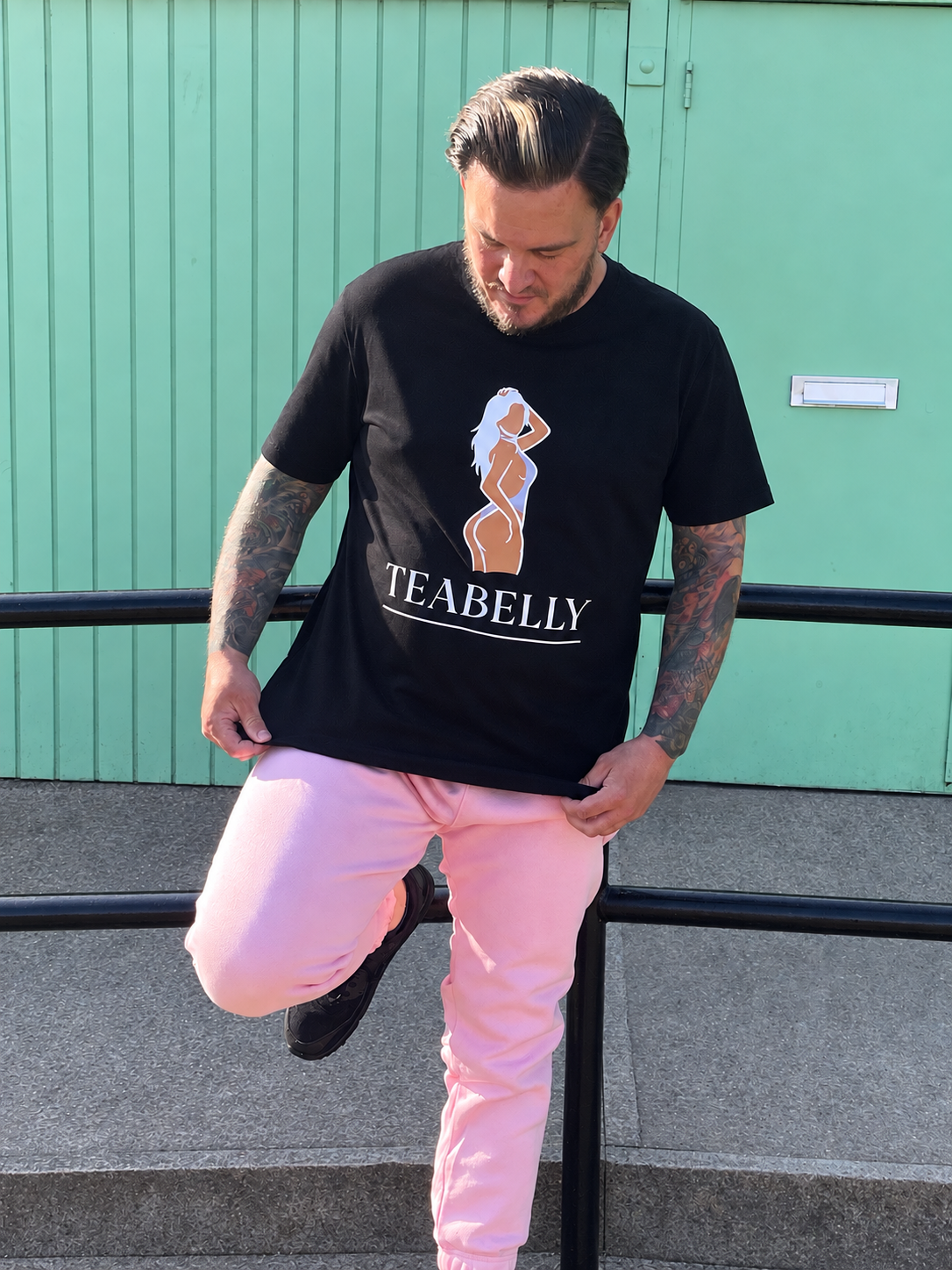 A man wearing a black graphic t-shirt with 'TEABELLY' and a woman's silhouette print, paired with light pink pants, posing outdoors against a mint green wall.